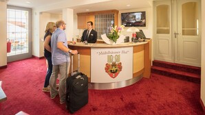 A hotel reception desk with a receptionist, a large window, and a red carpeted floor. - Hotel Mühlhäuser Hof (Mühlhausen/Thüringen)