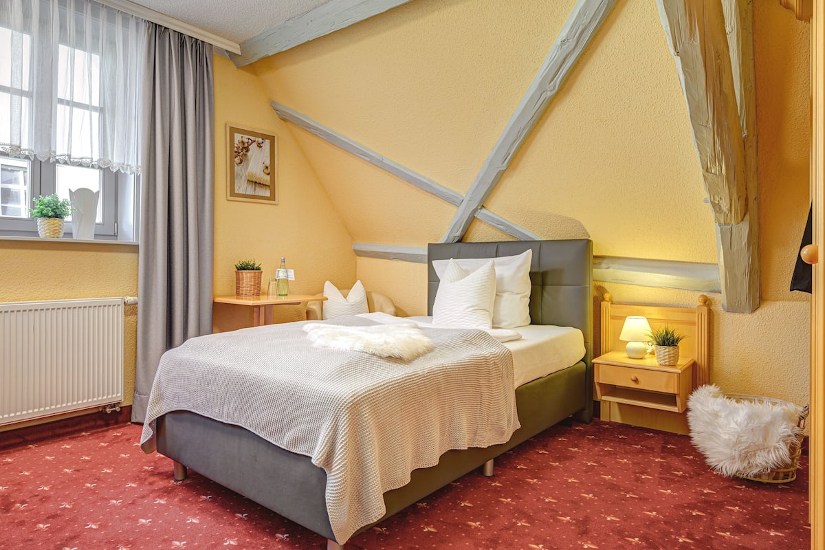 A bedroom with a bed, a nightstand, a lamp, a window with curtains, and a radiator.