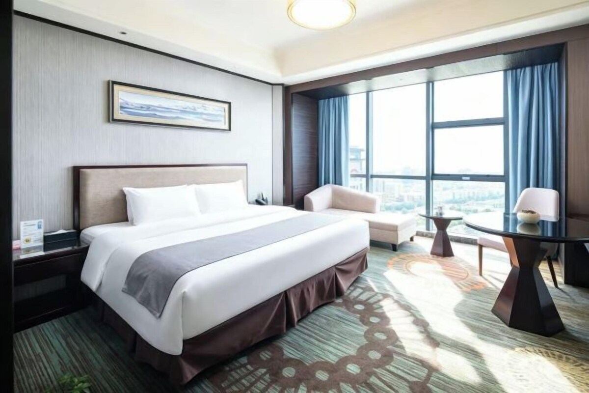 Habitación de hotel con una cama grande, una silla, una mesita y vistas a la ciudad a través de amplios ventanales.