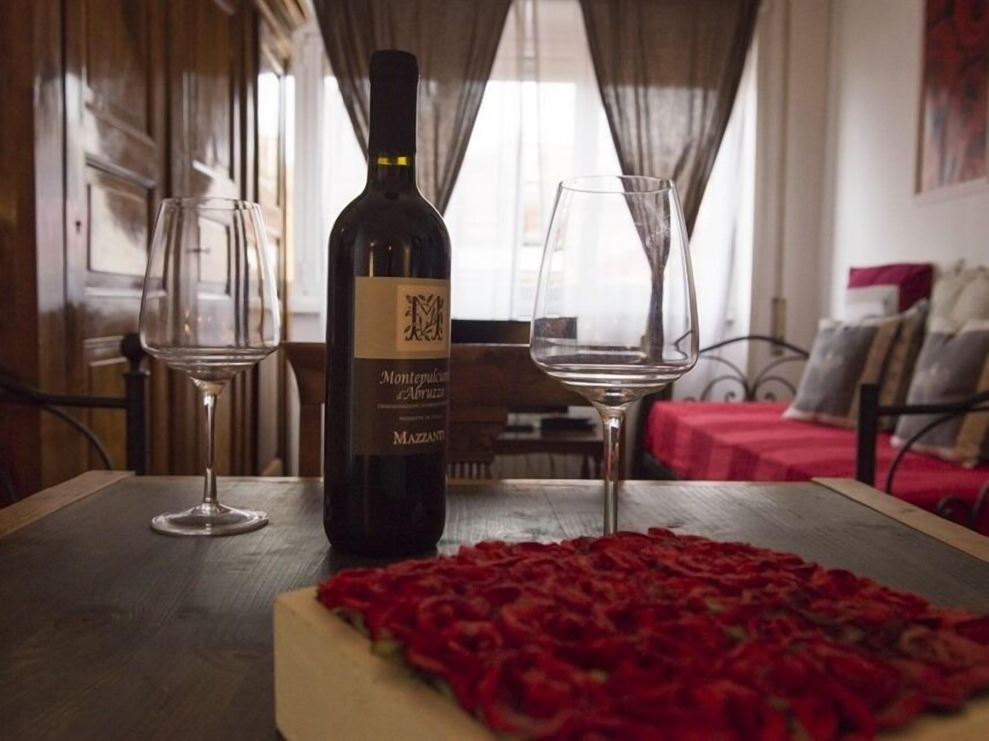 A bottle of Montepulciano d'Abruzzo wine, two empty wine glasses, and a red rose petal arrangement on a wooden table in a room with a red tablecloth and a bed with pillows in the background.