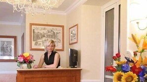 A reception desk with a woman seated behind it, framed black and white photographs on the wall, and a chandelier above. - AdoraSuite B&B (Rome)