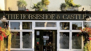 Un pub tradicional llamado "The Horseshoe & Castle" con una fachada clásica, ventanas amplias y plantas colgantes.
