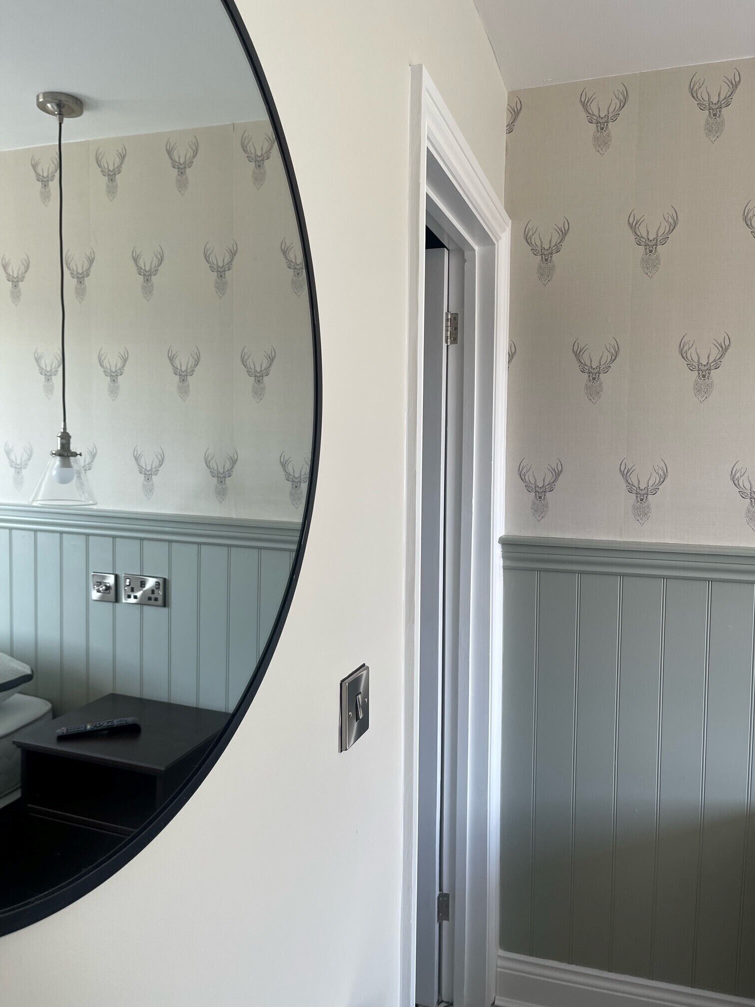 Une salle de bain avec un miroir rond, un lavabo et une porte donnant sur une autre pièce. Les murs sont recouverts d’un papier peint à motifs représentant des têtes de cerfs.