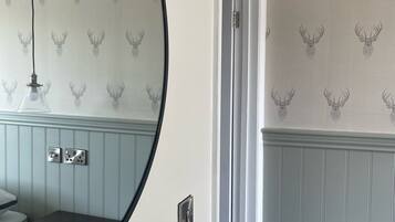Une salle de bain avec un miroir rond, un lavabo et une porte donnant sur une autre pièce. Les murs sont recouverts d’un papier peint à motifs représentant des têtes de cerfs.