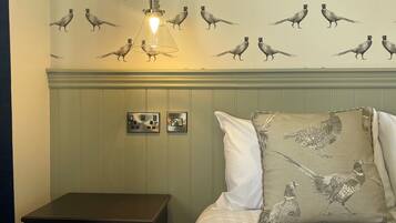 Een slaapkamer met behang met een vogelpatroon, een bed met wit beddengoed, een nachtkastje en een hanglamp.