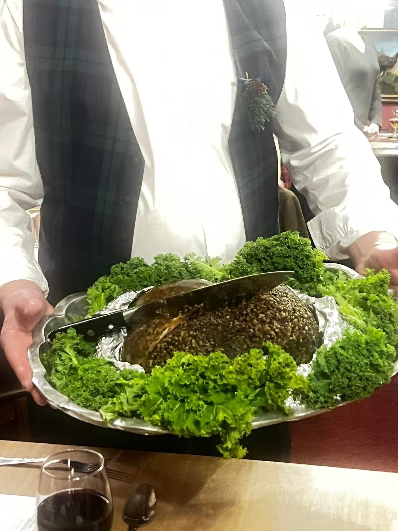 A person holding a large platter with a meat dish and greens, in a dining setting.