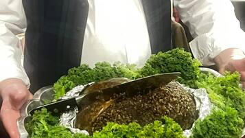 A person holding a large platter with a meat dish and greens, in a dining setting.