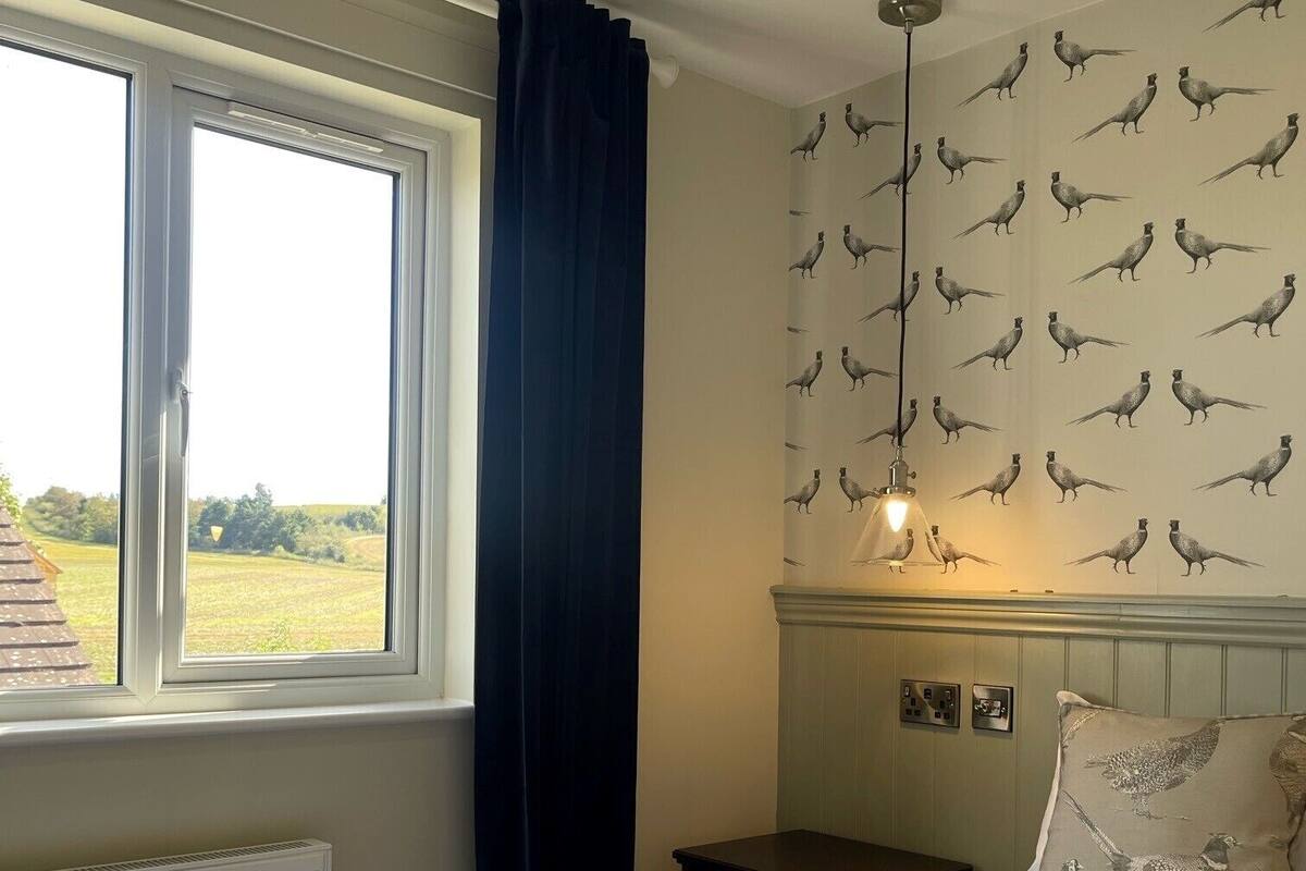 A bedroom with a large bed, a window with curtains, a wall with a bird pattern, a bedside table, and a hanging light fixture.