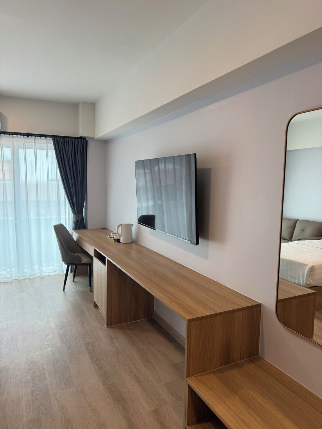 A modern hotel room with a wooden desk, a chair, a flat-screen TV mounted on the wall, and a large mirror reflecting the bed.