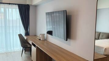 A modern hotel room with a wooden desk, a chair, a flat-screen TV mounted on the wall, and a large mirror reflecting the bed.