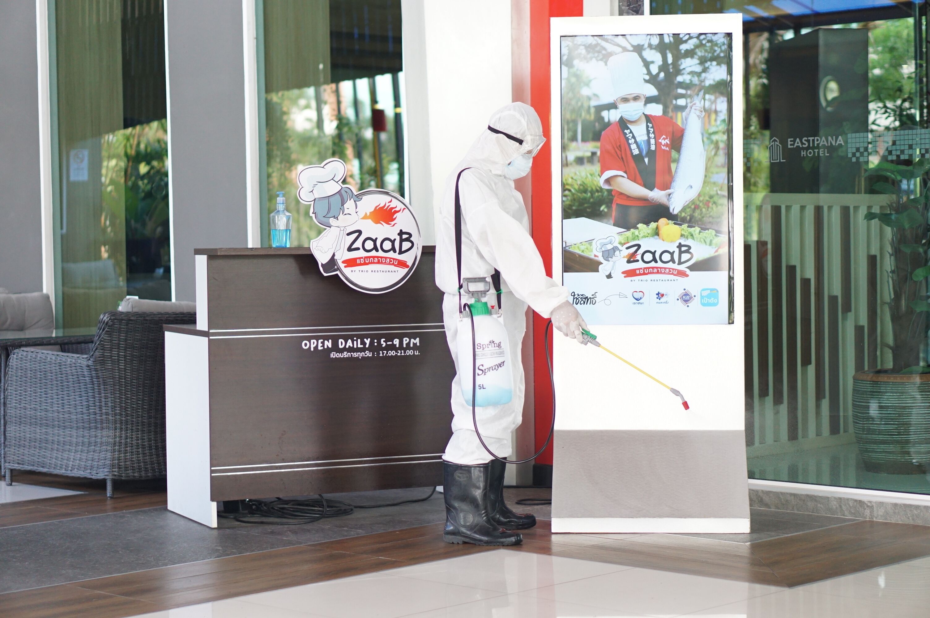 A person in a white protective suit spraying a liquid in a hotel lobby with a sign for 'ZaaB' and a 'Spaing Sprayer' bag.