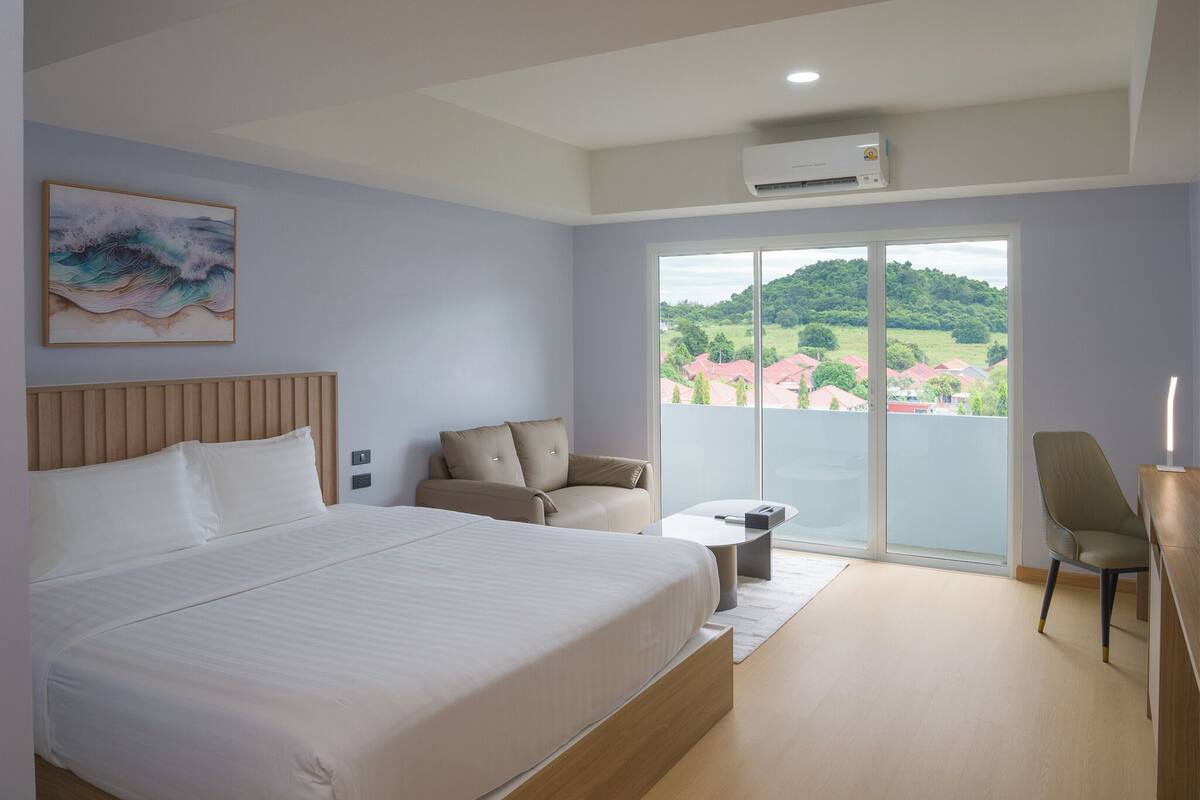 Een moderne hotelkamer met een groot bed, een bank, een bureau en een balkon met uitzicht op huizen en groen.