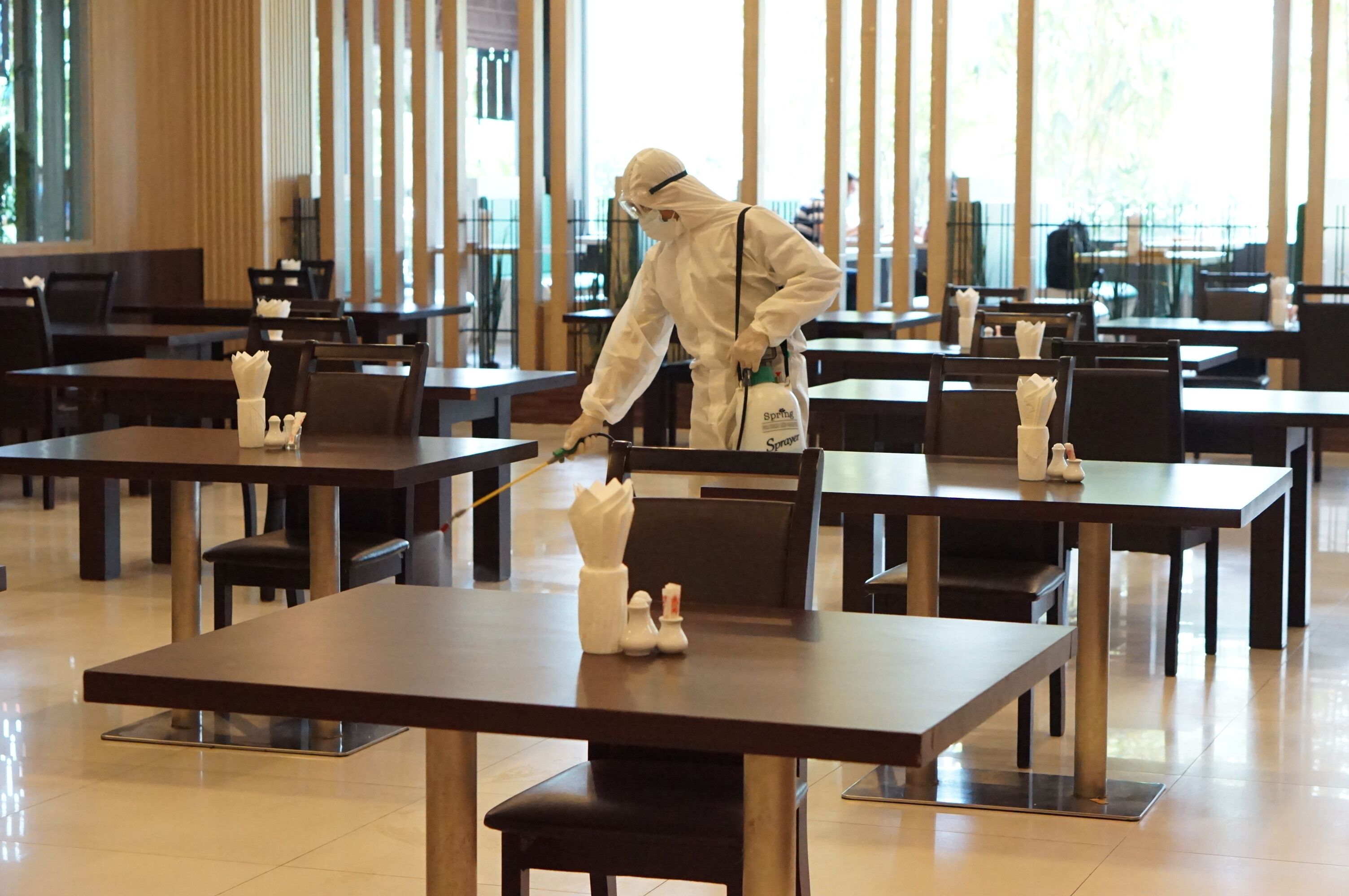 Une personne, vêtue d’une tenue de protection, nettoie des tables dans un grand espace de restauration bien éclairé, doté de mobilier moderne et de grandes fenêtres.