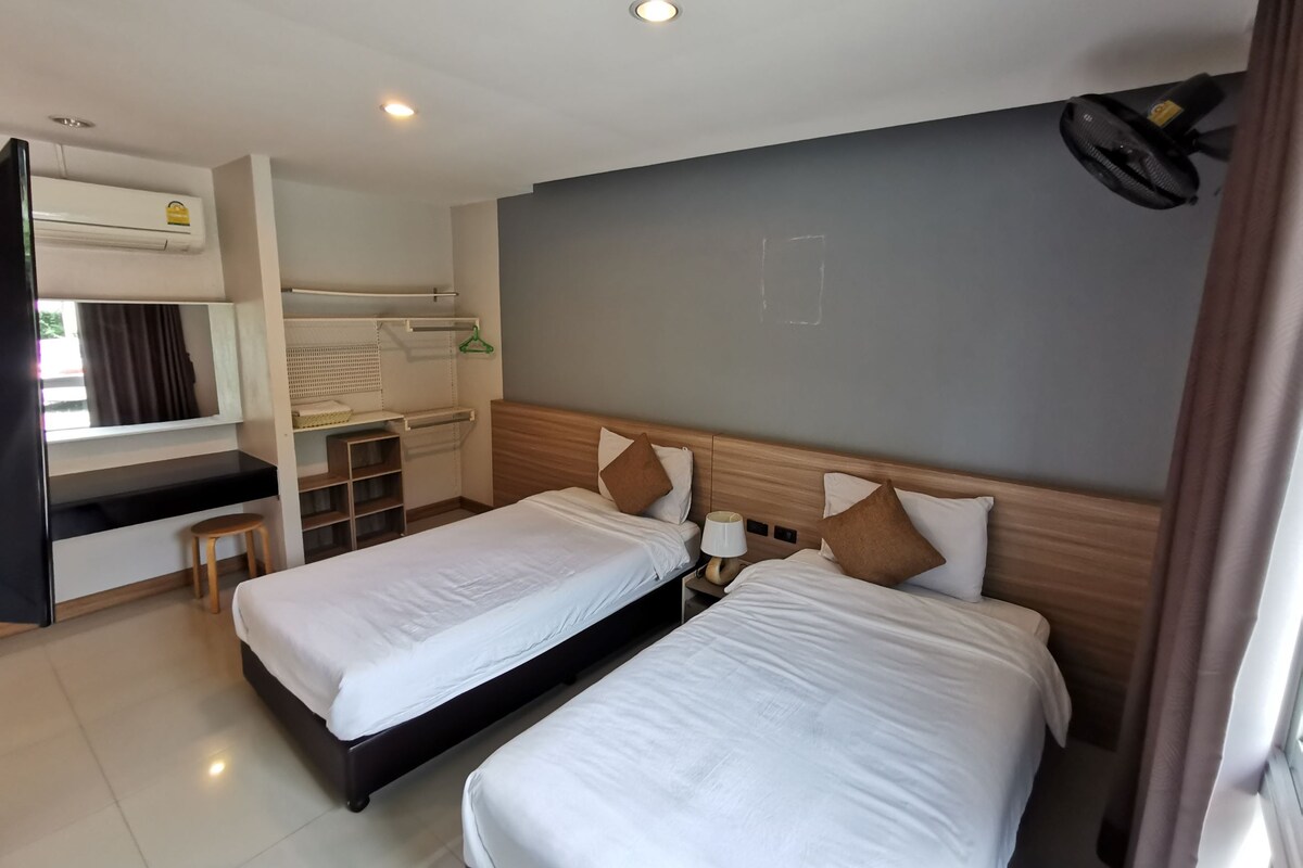 Quarto de hotel com duas camas, uma escrivaninha, uma cadeira, um espelho e um ventilador de teto.