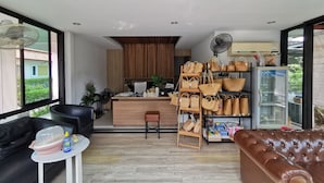 A modern living room with a black leather sofa, a brown leather couch, and a small white table. A kitchen area with wooden cabinets and a fan is visible in the background. - Smile Resort Sriracha (Si Racha)