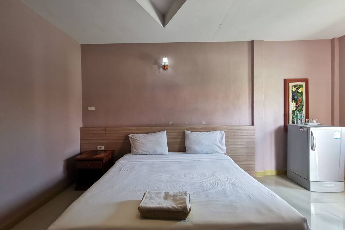 Hotellihuone, jossa on suuri sänky, yöpöytä, seinään kiinnitetty lamppu, jääkaappi ja seinällä oleva kehystetty kuva.