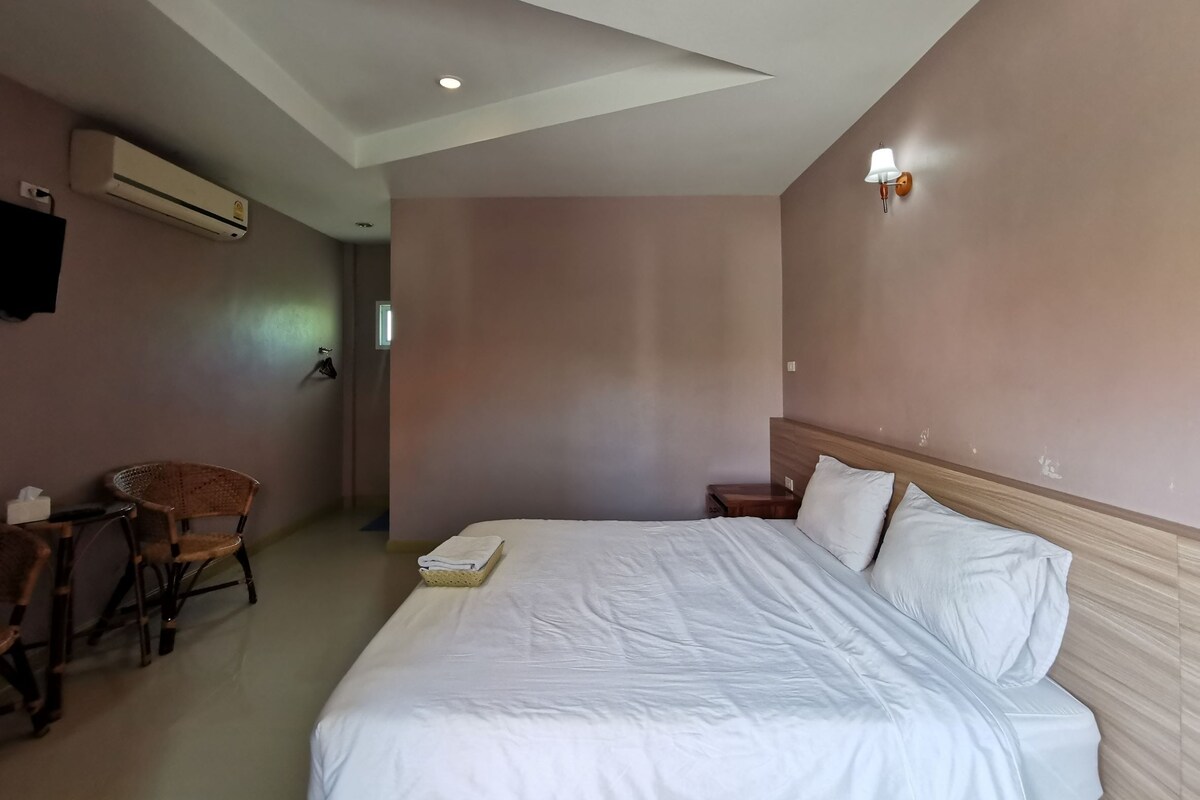 Quarto de hotel com uma cama grande, uma cadeira, uma mesinha e televisão fixada na parede.
