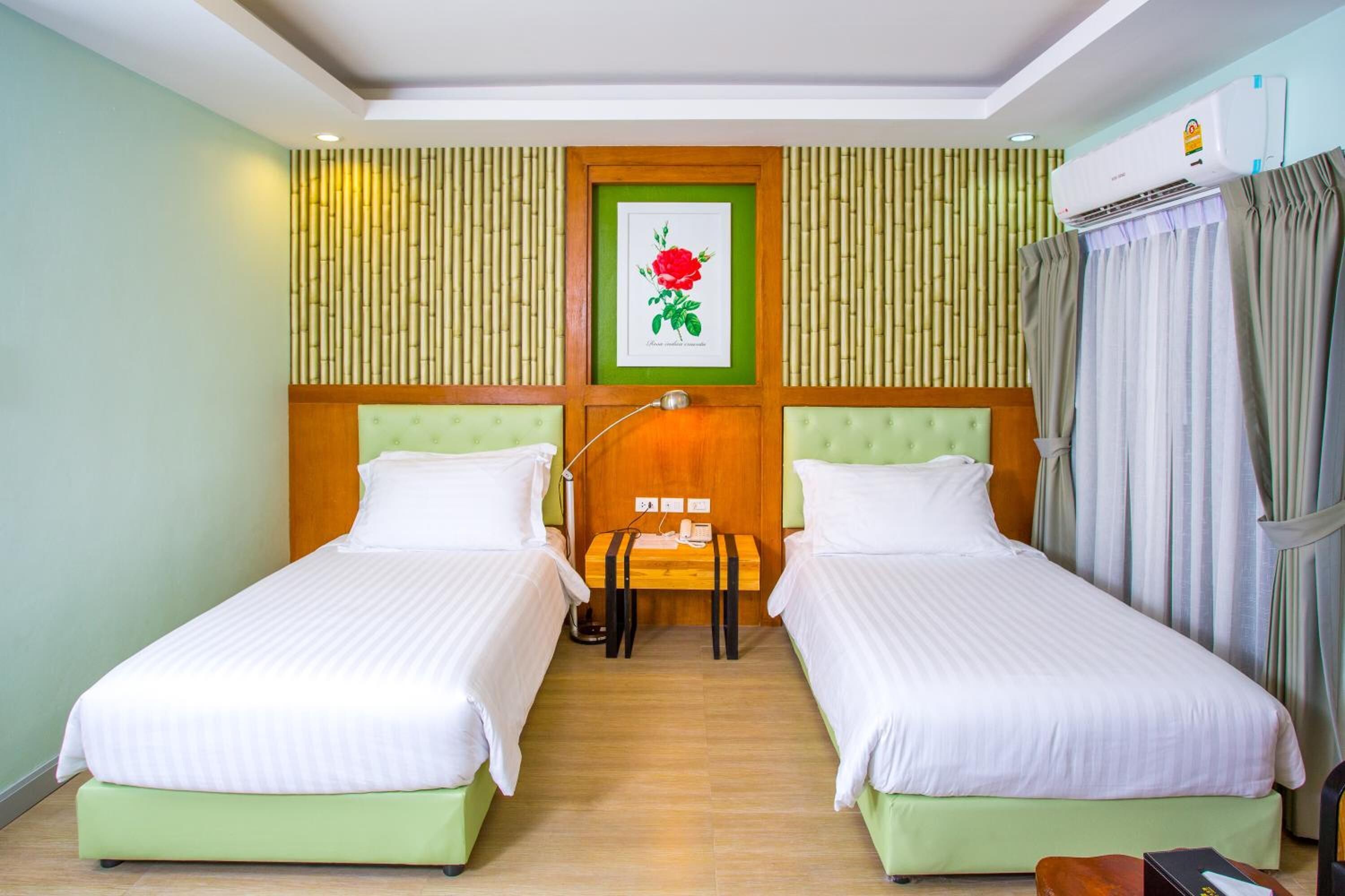 A hotel room with two single beds, a wooden headboard, a framed picture of a rose, a bedside table with a lamp, and an air conditioning unit.