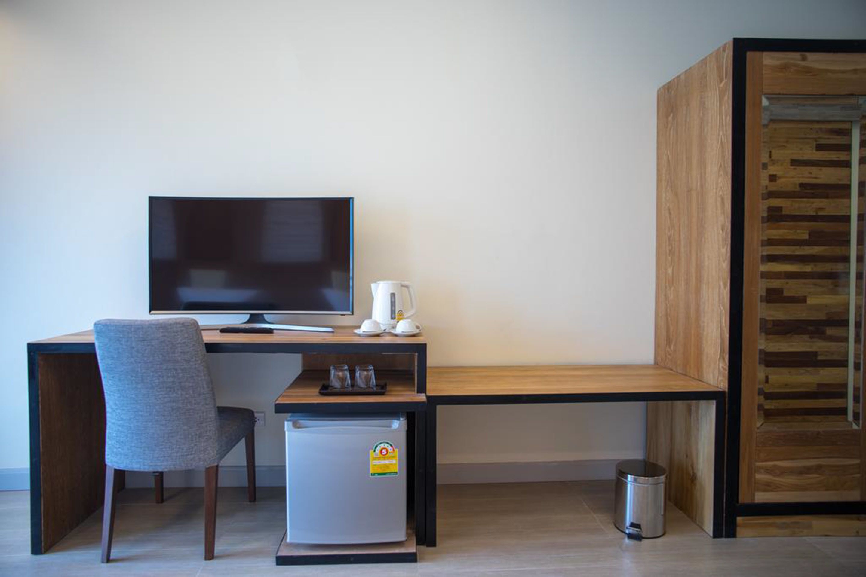 A compact room with a wooden desk, a television, a small refrigerator, a chair, a coffee maker, and a trash bin.