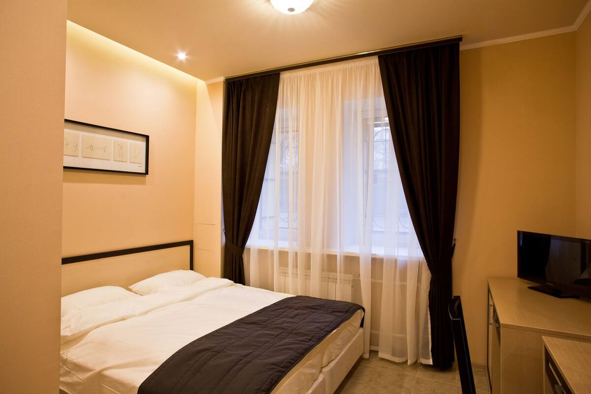 A hotel room with a bed, a television, and a window with sheer curtains.