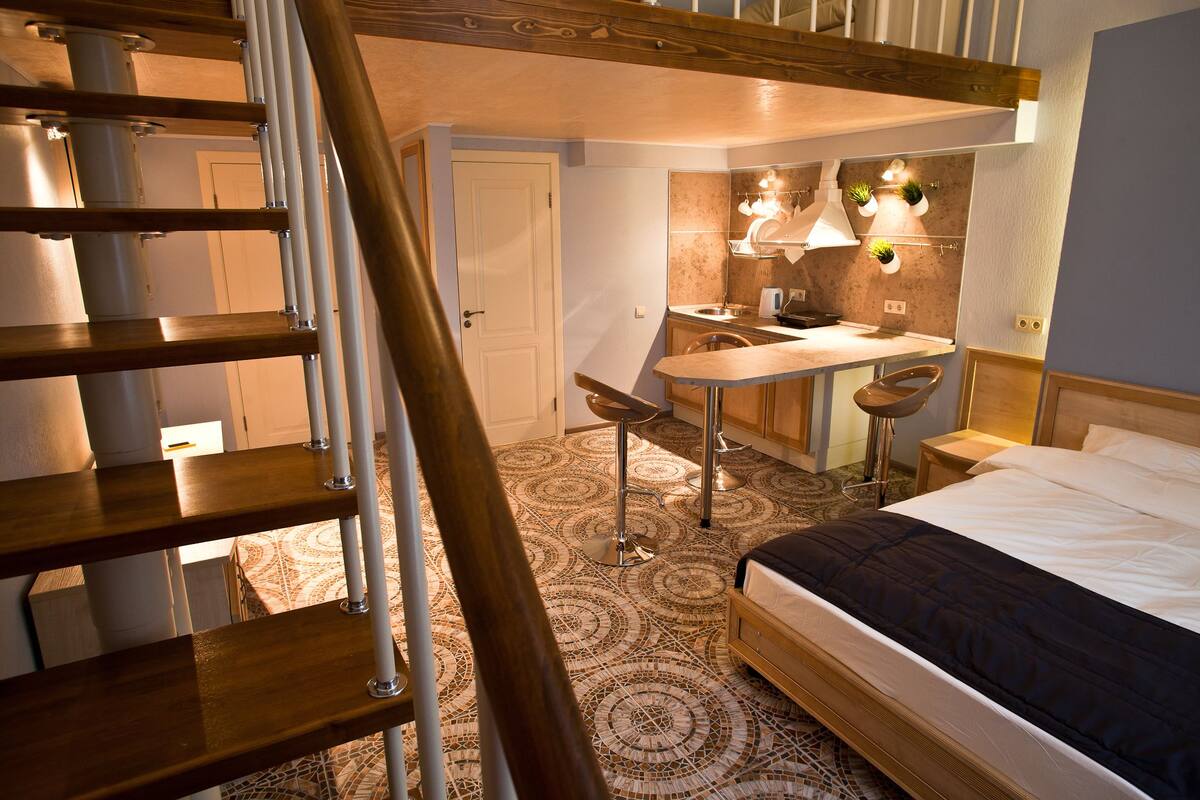 A bedroom with a bed, a wooden staircase, a kitchen area with a dining table and chairs, and a small bathroom.