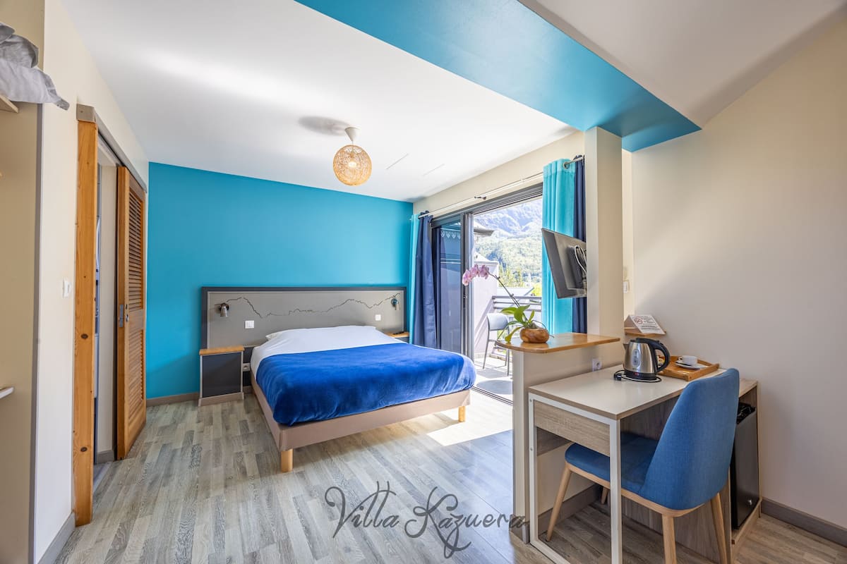 Une chambre à coucher avec un lit, un bureau et une chaise. Il y a une fenêtre avec des rideaux et une vue sur l’extérieur.