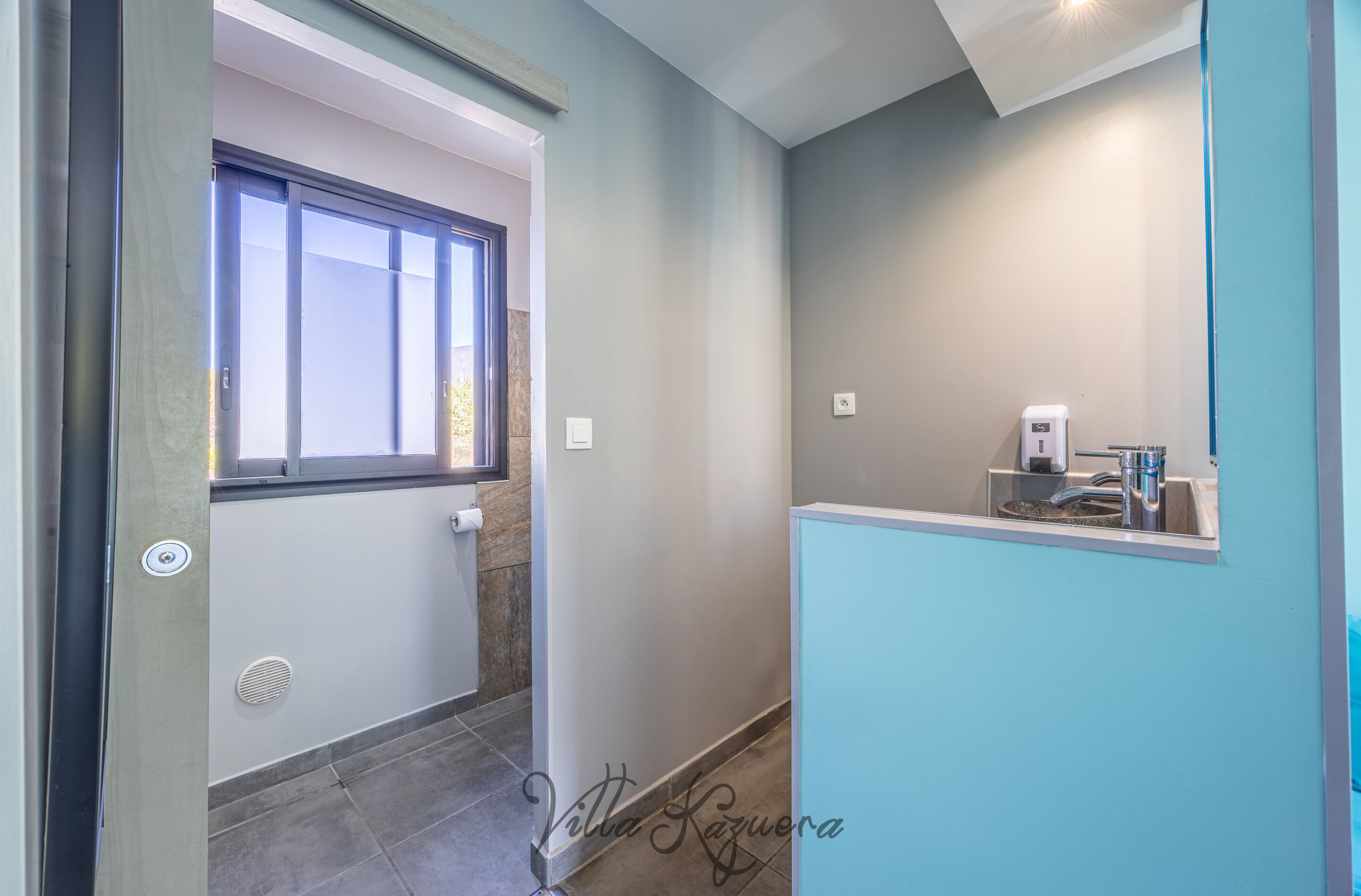 A bathroom with a window, a sink, and a towel dispenser.