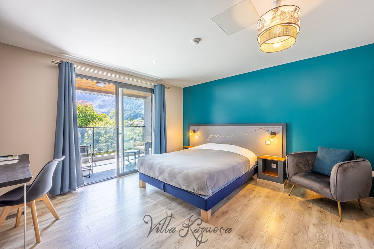 Une chambre moderne avec un lit, un bureau et une chaise. Elle dispose d’un balcon avec vue sur les montagnes.