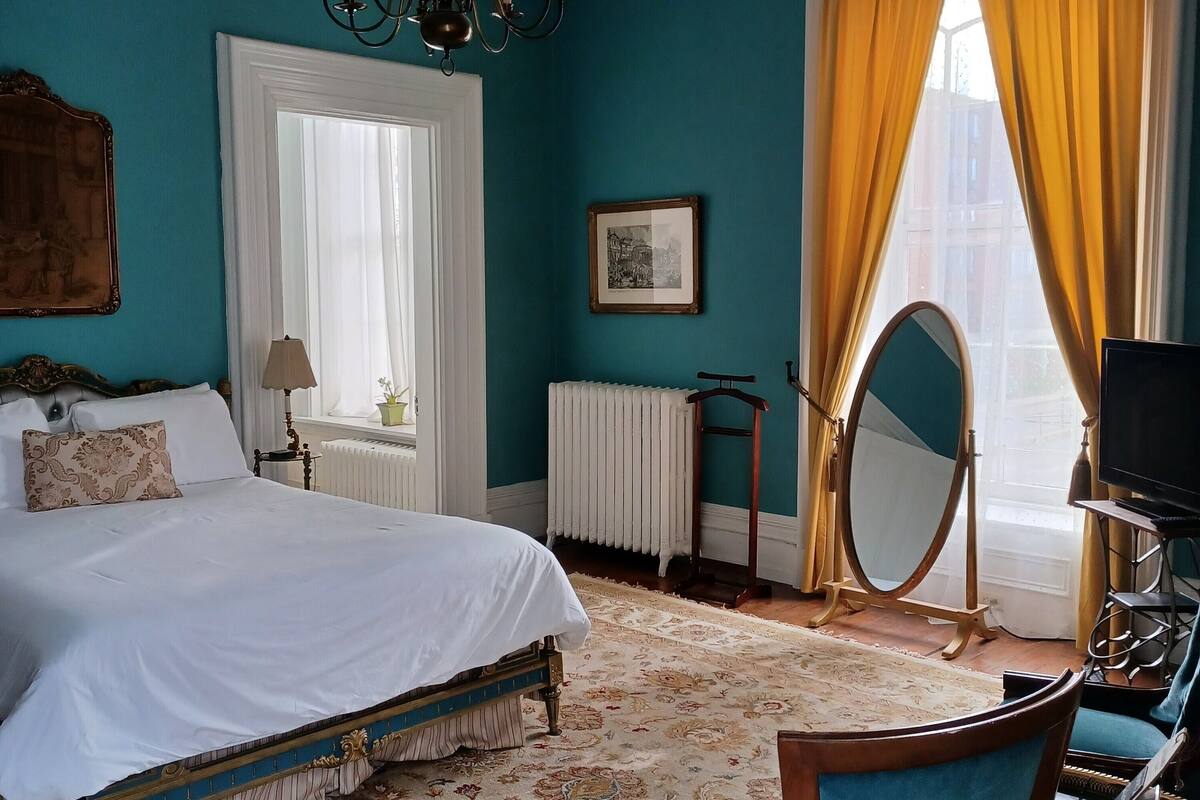 A bedroom with a large bed, a chandelier, a window with curtains, a radiator, a mirror, and a television.