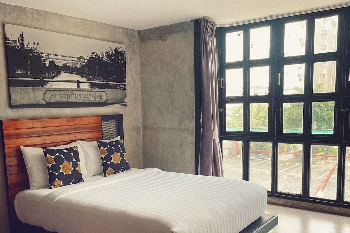 Een moderne slaapkamer met een groot bed, een houten hoofdbord, een stadslandschap schilderij en uitzicht op een straat door een groot raam.