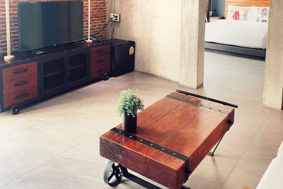 Een moderne woonkamer met een bakstenen muur, een grote flatscreen-tv, een houten salontafel met wielen, en een bed op de achtergrond.