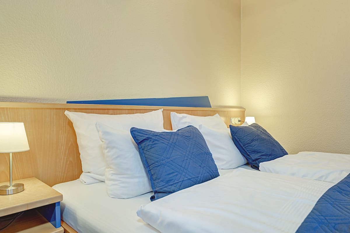 A hotel room with a bed, white bedding, a blue quilted pillow, a bedside lamp, and a wooden headboard.