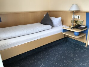 A single bed with a white mattress and a grey pillow, a bedside table with a lamp, a telephone, and a radio.
