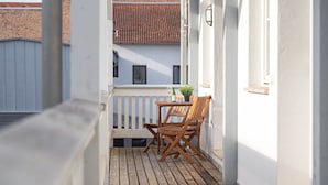 Una terraza de madera con una mesa y sillas, una planta en maceta y vista a un edificio con techo de tejas rojas.