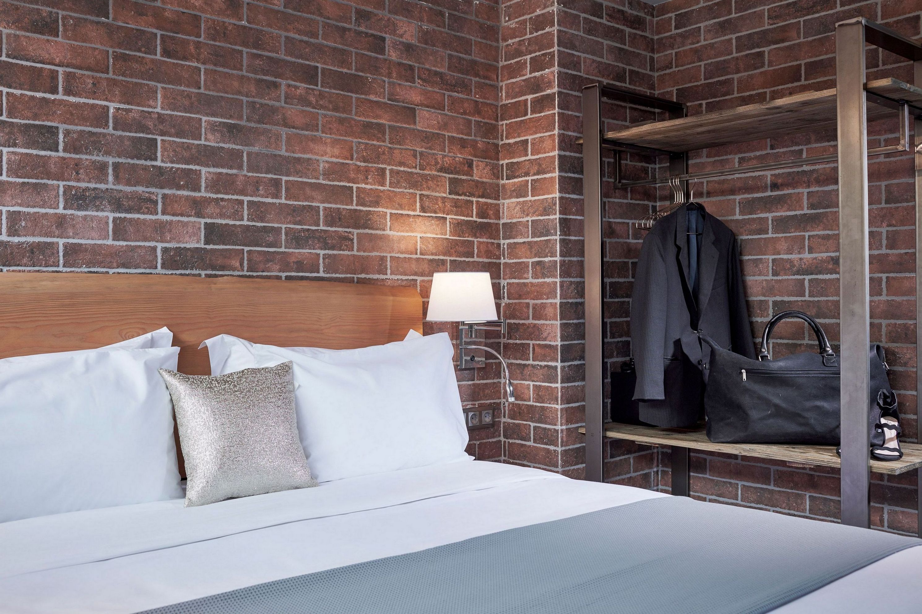 Quarto de hotel com parede de tijolos, cabeceira de madeira, cama com roupa de cama branca, almofada prateada, bolsa de mão preta e um casaco numa prateleira.