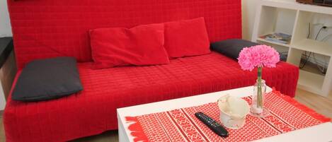 A red sofa with black and red cushions, a white coffee table with a red placemat, a remote control, a vase with pink flowers, and a bookshelf with books and a radio.