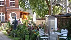 A garden patio with outdoor seating, a statue, and a brick building in the background. - Ferienwohnung Backstube König (Brandenburg an der Havel)