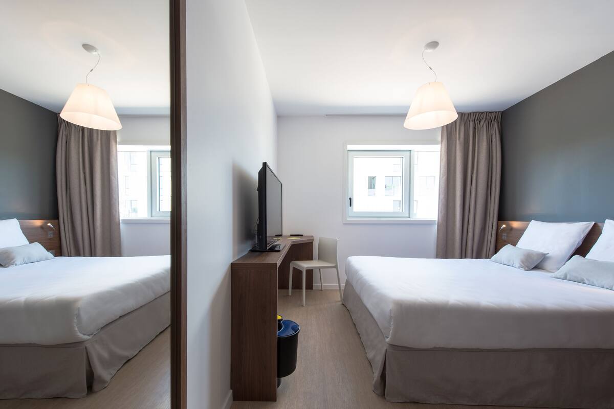 A hotel room with two beds, a desk, a chair, a TV, and a window.