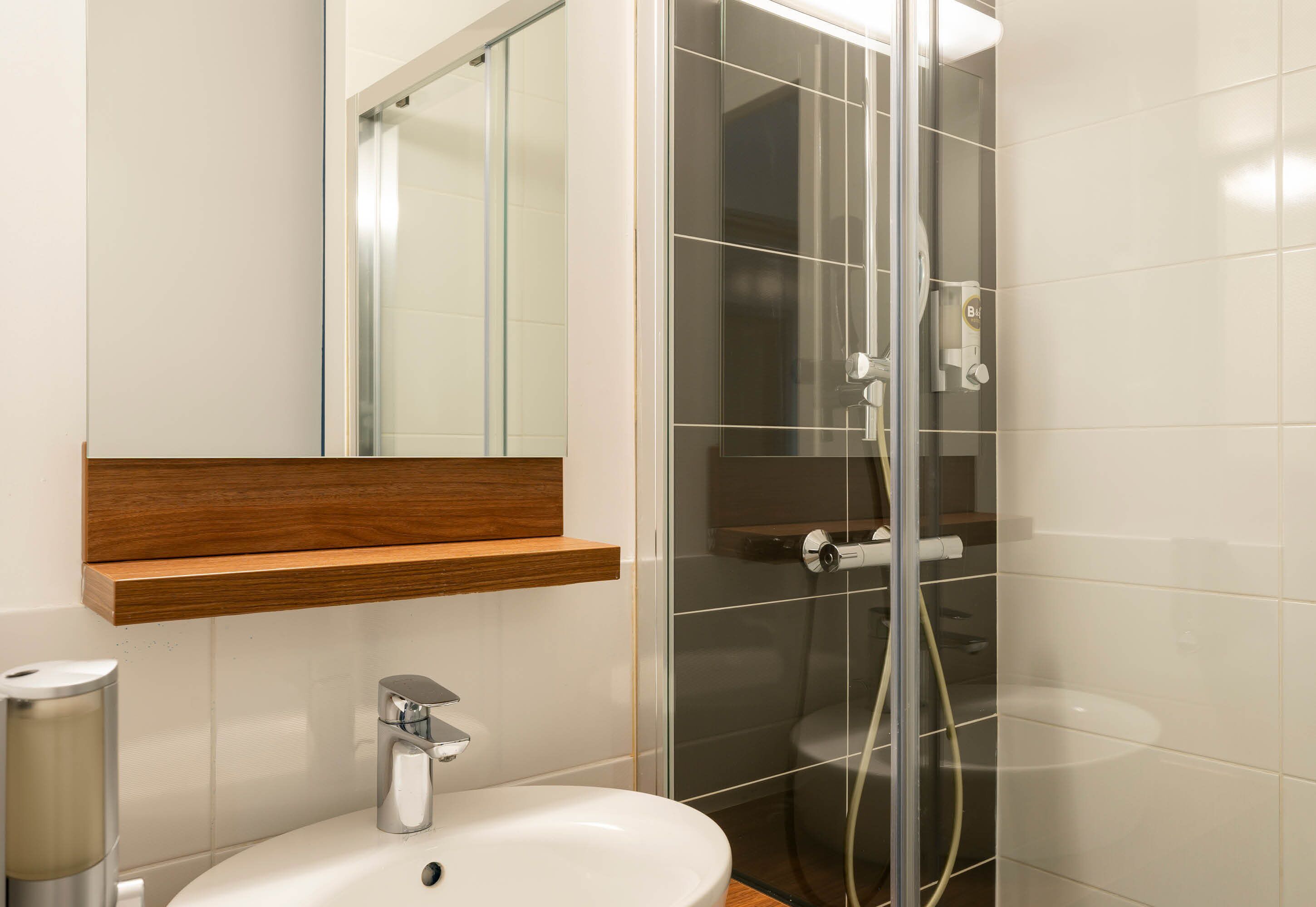 A modern bathroom with a glass shower enclosure, a white sink, and a wooden shelf above the mirror.