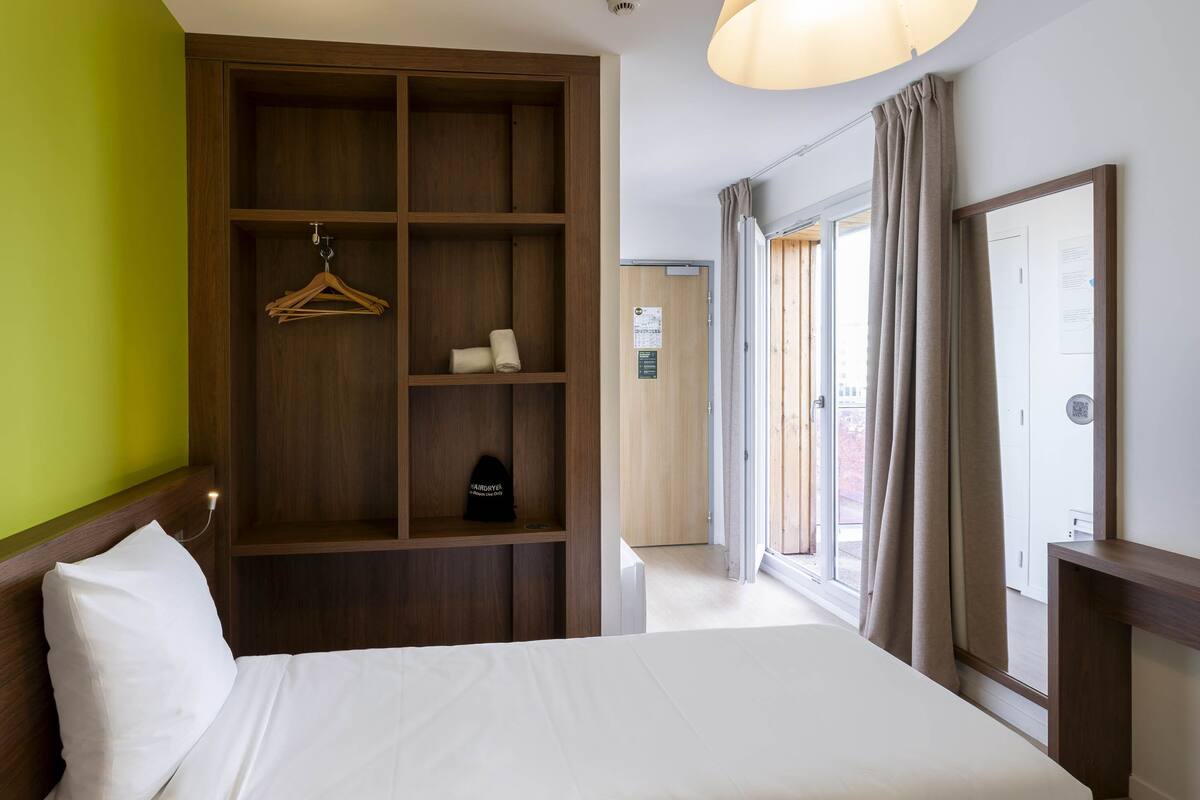 A hotel room with a large bed, a wardrobe with multiple shelves, a hanging coat rack, and a view of the outside through a window with curtains.