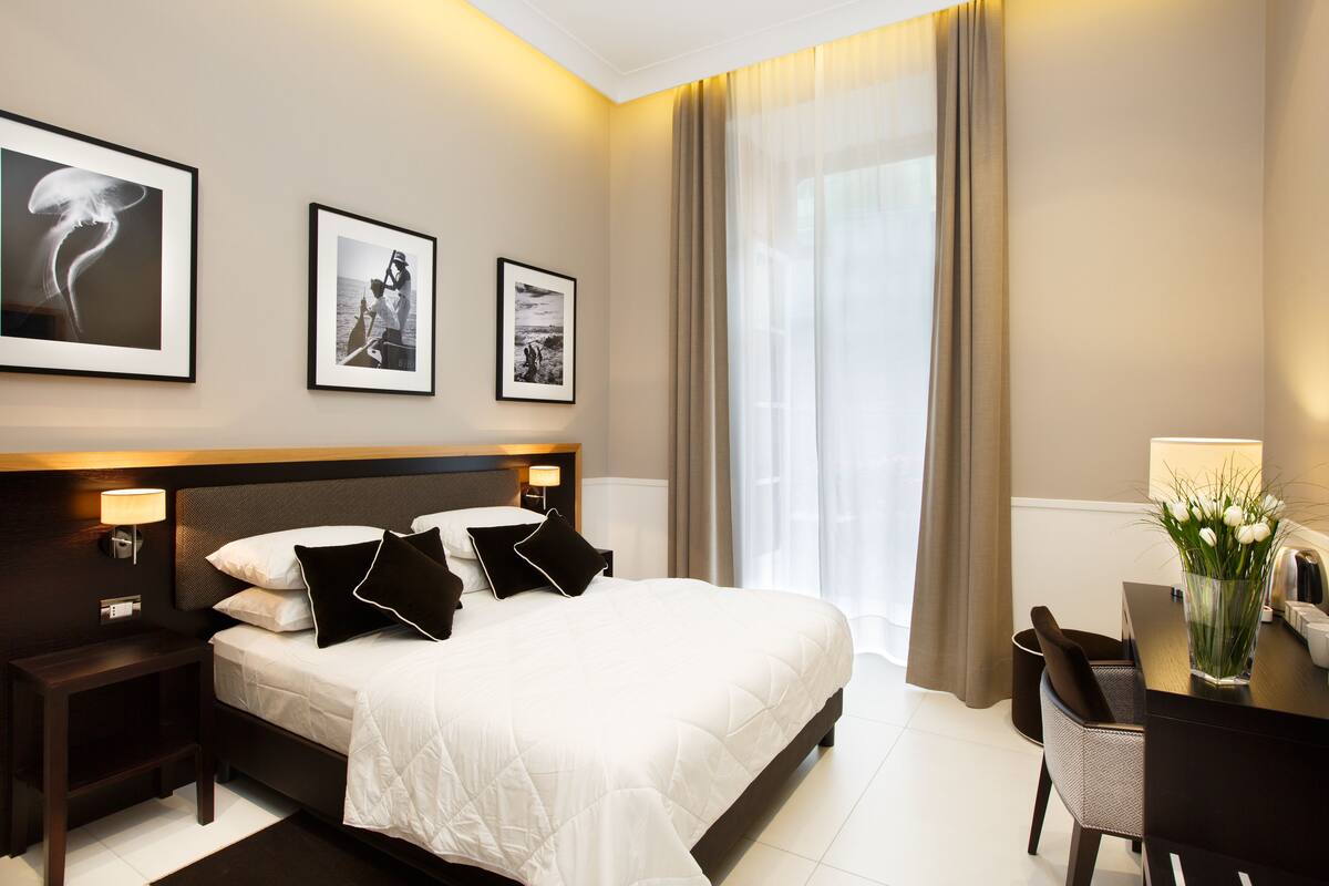 Une chambre d’hôtel moderne avec un grand lit, des tables de chevet, un bureau et une chaise. Des tableaux encadrés ornent le mur, et un vase rempli de fleurs est posé sur le bureau.