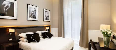 A modern hotel room with a large bed, bedside tables, a desk, and a chair. There are framed pictures on the wall and a vase with flowers on the desk.