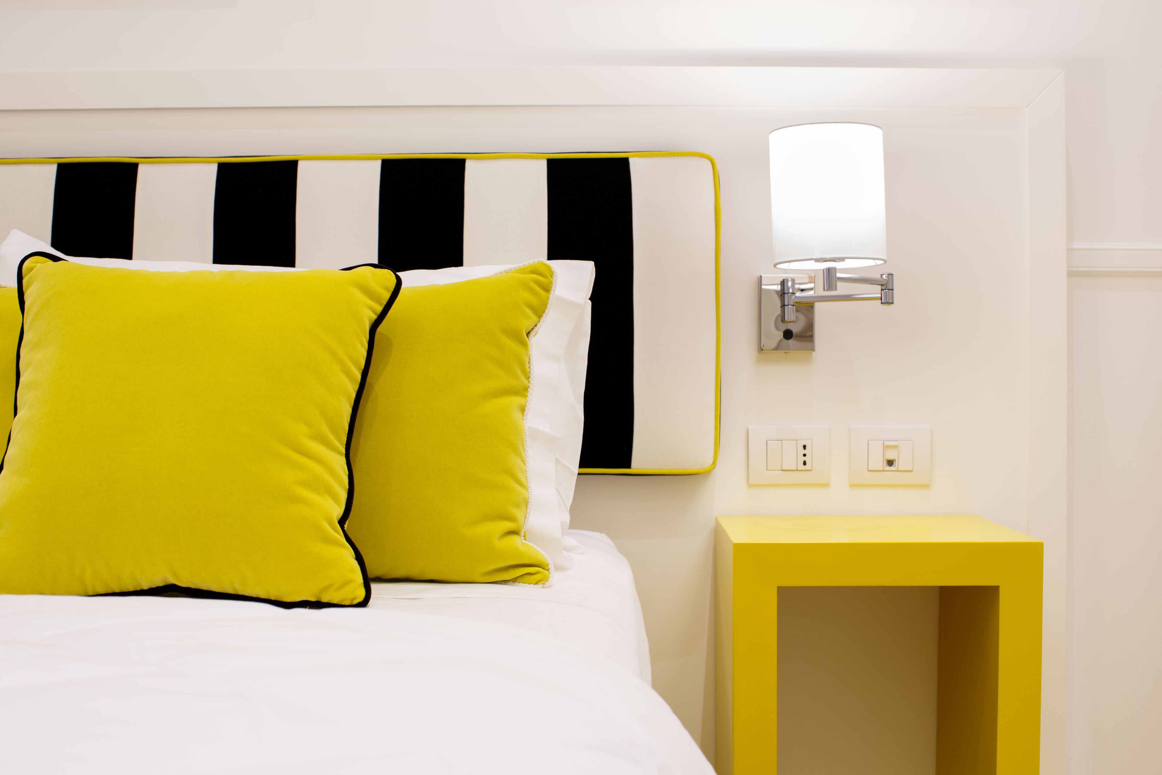 Een bed met een hoofdbord met witte en zwarte strepen, twee heldergele kussens, een nachtkastje en een wandlamp.