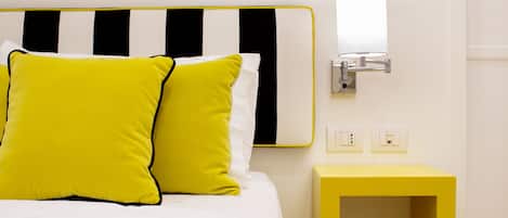 Ein Bett mit weiß-schwarz gestreifter Kopfteil, zwei leuchtend gelbe Kissen, ein Nachttisch und eine wandmontierte Lampe.