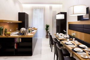 Un comedor moderno con una larga mesa de madera dispuesta para una comida, una zona de cocina con mostrador, y una sala de estar con sofá y sillones.