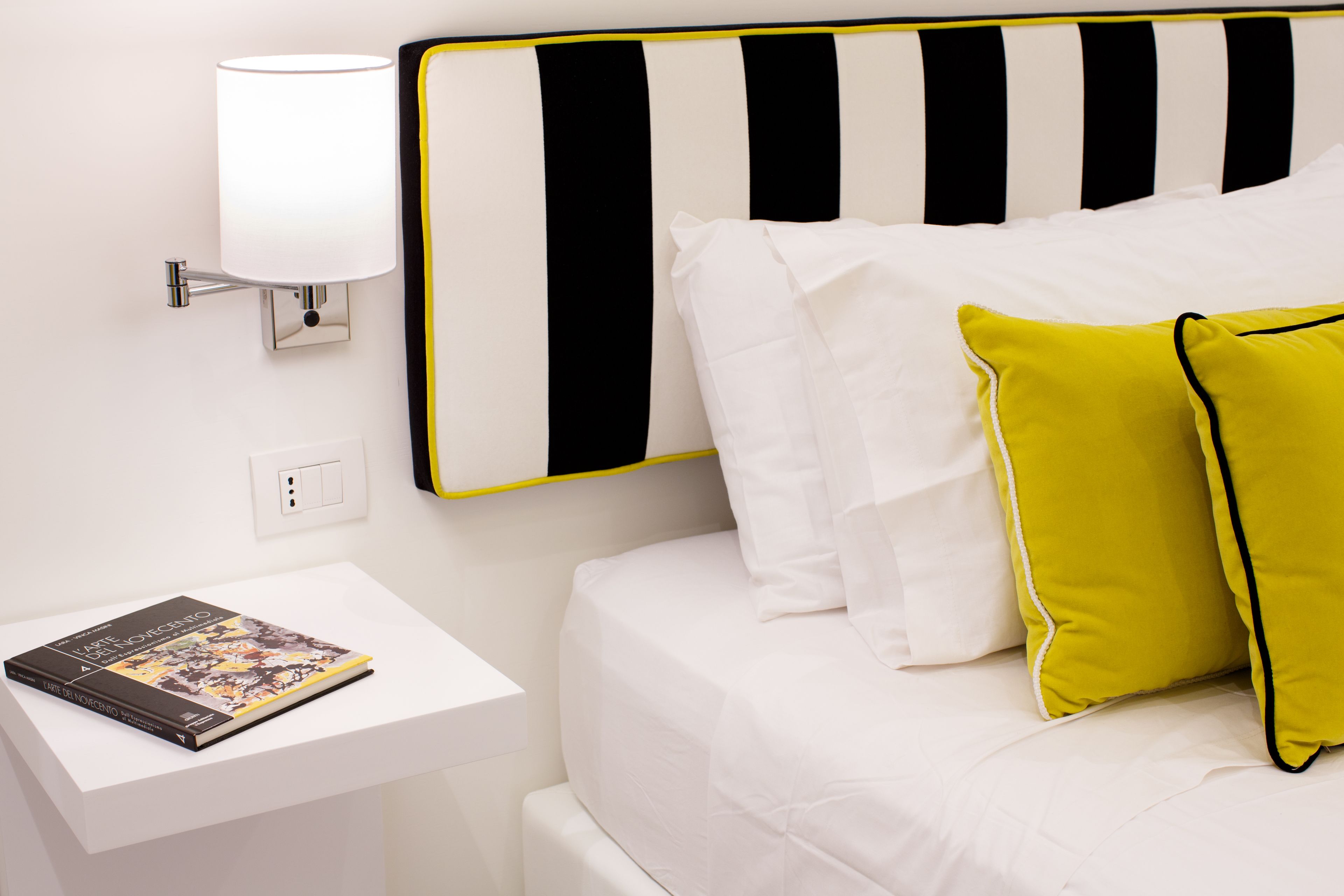 A neatly made bed with white and yellow pillows, a black and white striped headboard, and a bedside table with a lamp and a book.