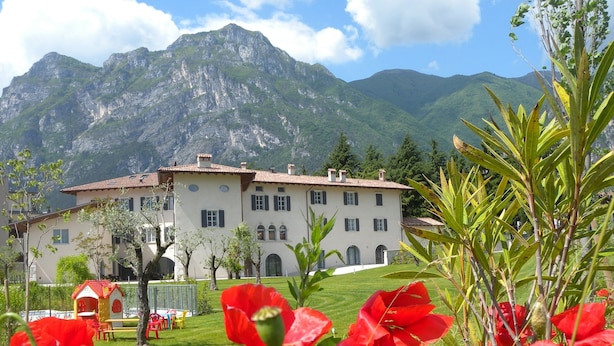 A large building with a white facade and red-tiled roof, surrounded by lush greenery and mountains in the background. - Borgo San Nazzaro (Riva del Garda)