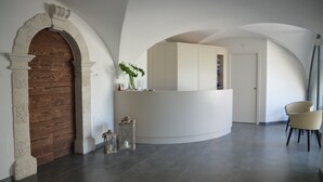 A reception area with a wooden door, a white reception desk, a vase with flowers, and a chair. - Borgo San Nazzaro (Riva del Garda)