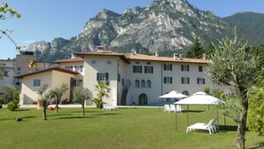 A large building with multiple windows and a staircase leading to the entrance. There are outdoor seating areas with umbrellas and lounge chairs. - Borgo San Nazzaro (Riva del Garda)
