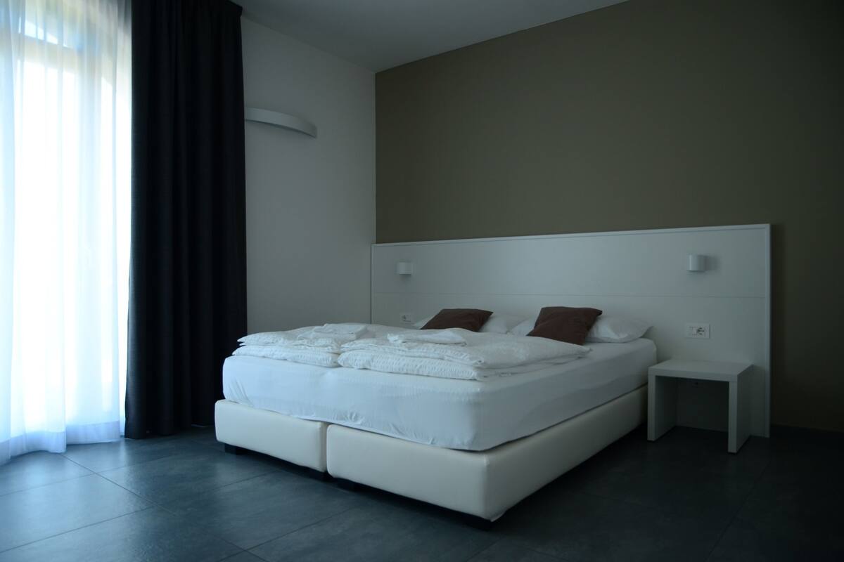 Ein modernes Schlafzimmer mit einem großen Bett, einem Nachttisch und Blick ins Freie durch transparente Vorhänge.